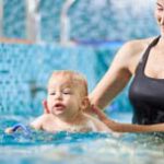 Young smiling mother is helping her little son swim in turquoise water during swimming class for infants, baby is splashing around in water. Concept of active family leisure